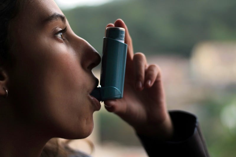 A female presenting person holding up an inhaler and taking a puff